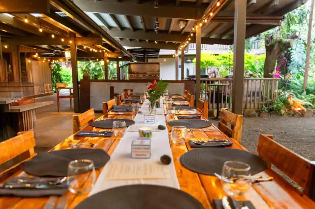 Long wooden dining table set for a group meal beneath string lights in a covered tropical outdoor venue.