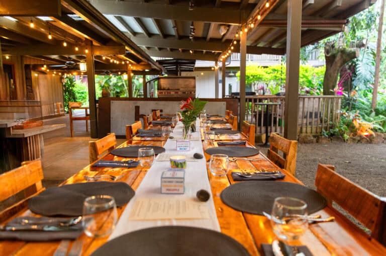 Long wooden dining table set for a group meal beneath string lights in a covered tropical outdoor venue.