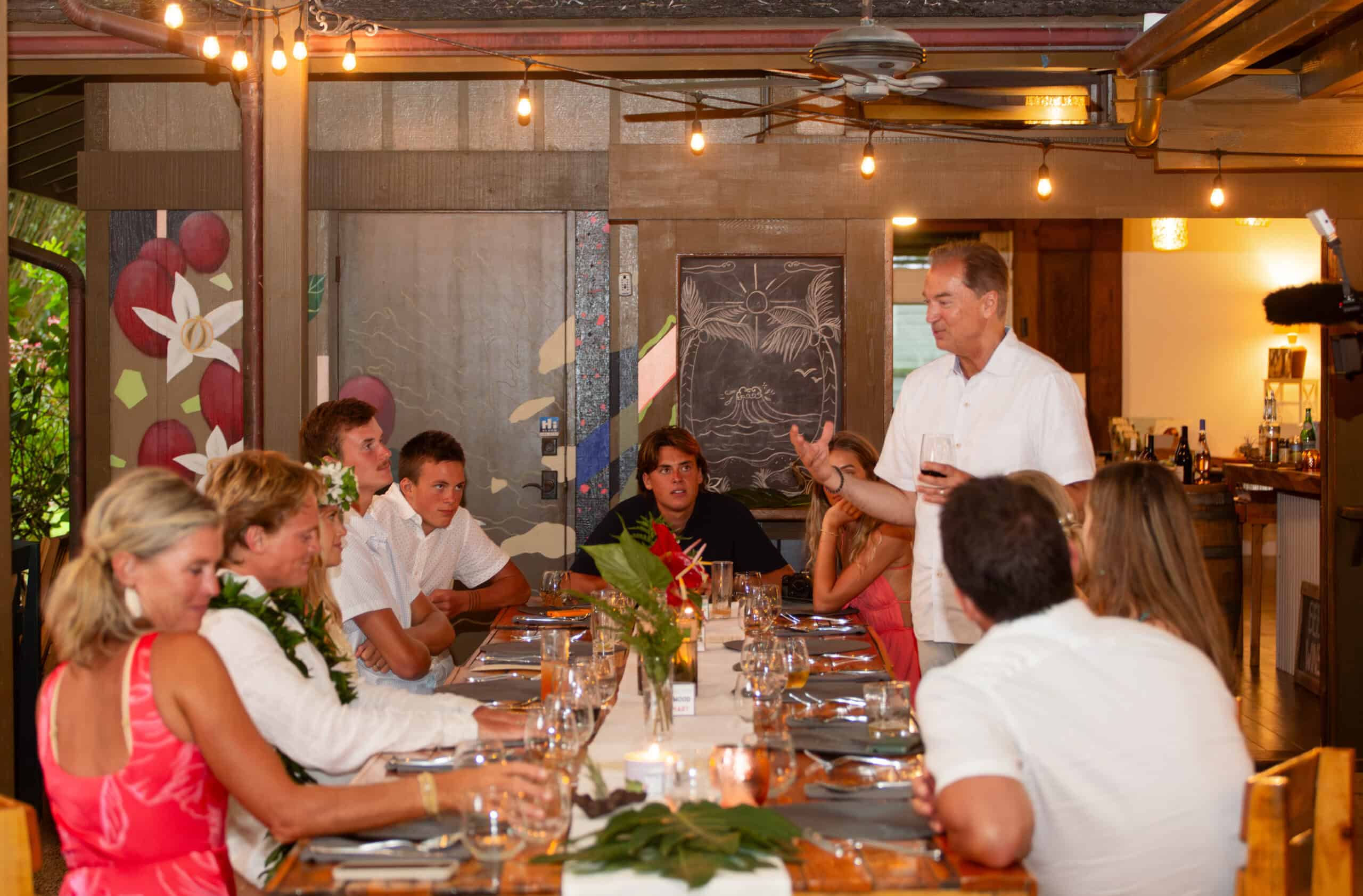 Man giving a toast to guests seated at a long communal dining table during an open-air event in Kauaʻi.
