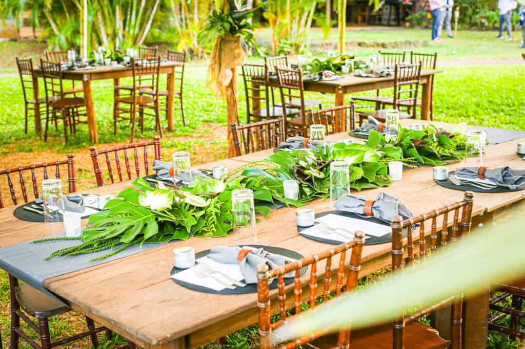 Outdoor dining setup with wooden tables, tropical greenery centerpieces, and place settings on a sunny lawn.