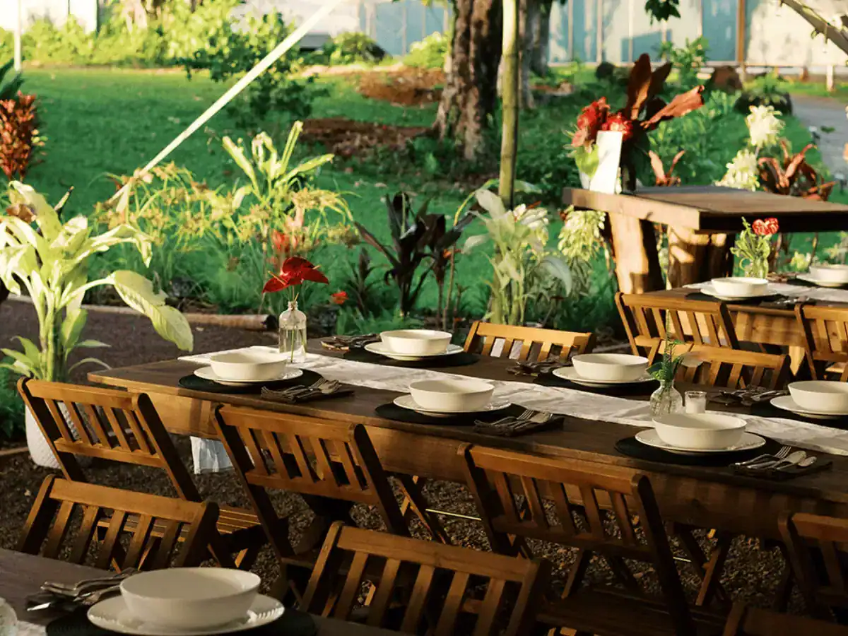 Long wooden tables set with white bowls, black place settings, and floral accents in a tropical outdoor garden dining area.