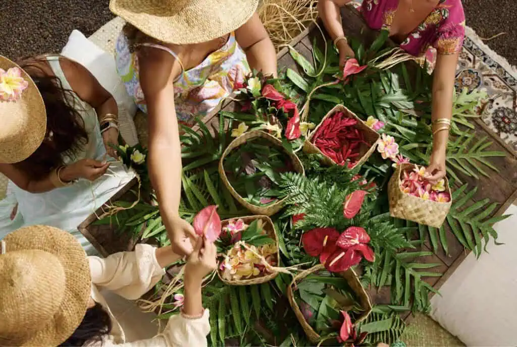 Overhead view of guests weaving tropical flowers and greenery into colorful Hawaiian lei around a wooden table.